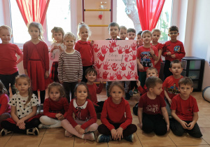 Dzieci z grup: Słoneczka i Promyczki, ubrane w czerwone bluzki, trzymają plakat, na którym są czerwone odciski dłoni wycięte z papieru i napis Stop przemocy!
