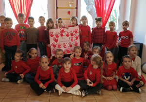 Dzieci 6-letnie,ubrane w czerwone bluzki, trzymają plakat, na którym są czerwone odciski dłoni wycięte z papieru i napis Stop przemocy!