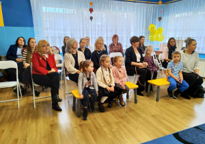 Dzieci siedzą na ławeczkach, za nimi wiceprezydent Małgorzata Kańska-Kipigroch, dyrektorzy przedszkoli i opiekunowie dzieci.
