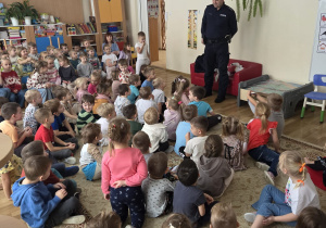 Przedszkolaki siedzą na podłodze i słuchają pana policjanta.
