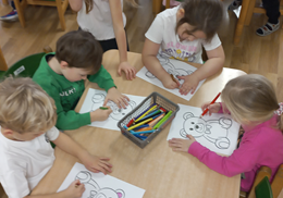 Przedszkolaki siedzą przy stolikach i kolorują sylwetę misia.