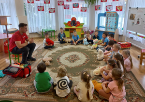 Dzieci siedzą w kole na dywanie z pluszowymi misiami. Przed nimi siedzi ratownik medyczny.