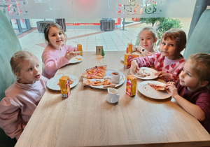 Pięć dziewczynek siedzi przy stoliku, jedzą pizzę.