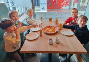 Pięciu chłopców siedzi przy stoliku i pokazują jaką smakowitą pizzę wykonali.