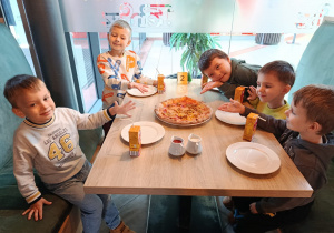 Pięciu chłopców siedzi przy stoliku i pokazują jaką smakowitą pizzę wykonali.