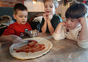 Chłopiec smaruje placek pizzy sosem pomidorowym. Dwóch chłopców się przygląda.