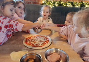 Dziewczynki sypią starty ser na placek pizzy posmarowany sosem pomidorowym.