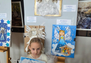 Maja z dyplomem w rękach na tle tablicy z nagrodzonymi pracami.