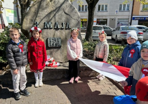 Dzieci stoją pod pomnikiem z rozłożoną flagą.